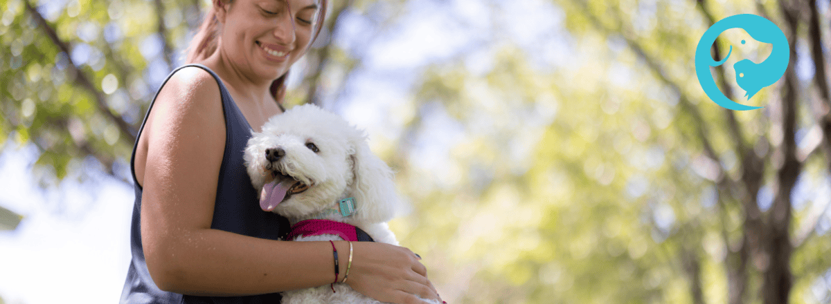 A woman smiling outdoors with her fluffy white dog climbing into her lap. Dog listens to one partner but not the other.