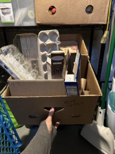 A hand pulling a large cardboard box off a shelf to see inside. It is full of other cardboard and plastic materials. There is writing on the box that says "Pete Trash."