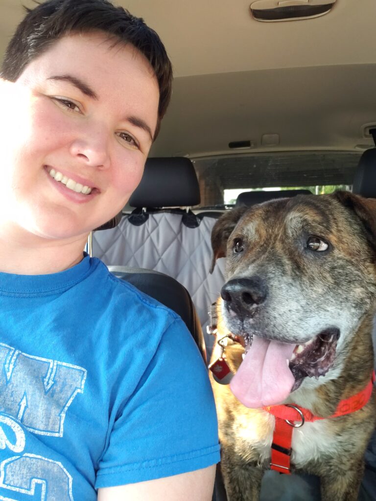 A woman with short dark hair sitting next to a brindle dog with a grey muzzle in a car.