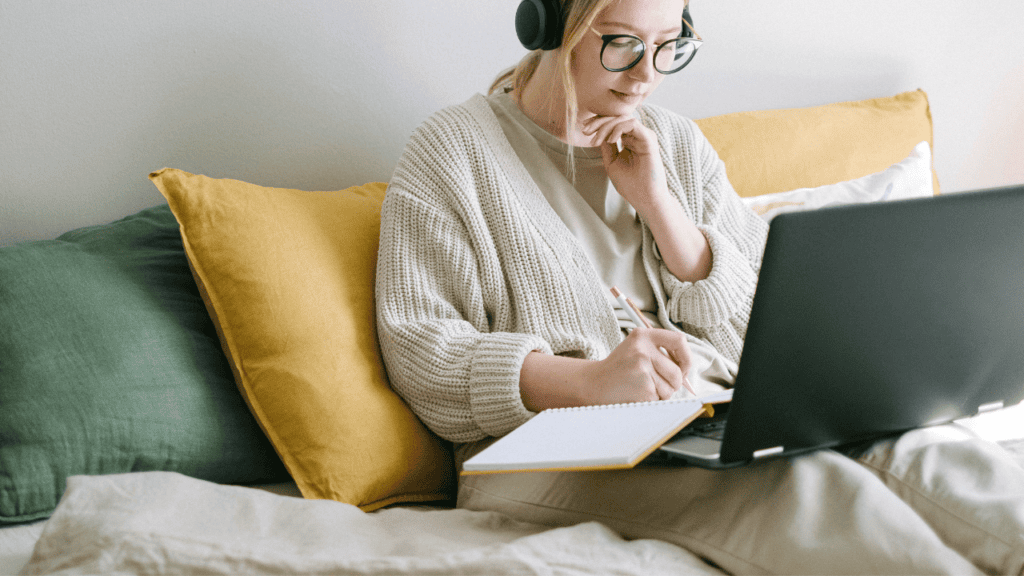 A woman sitting on a couch with colorful pillows with headphones and a laptop taking a webinar.