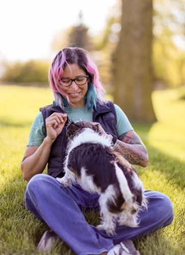 A woman with colorful hair sitting in the grass with a small scruffy dog on her lap, feeding her treats. Veronica Garcia, CVT and Fear Free certified senior dog behavior consultant at Pet Harmony.