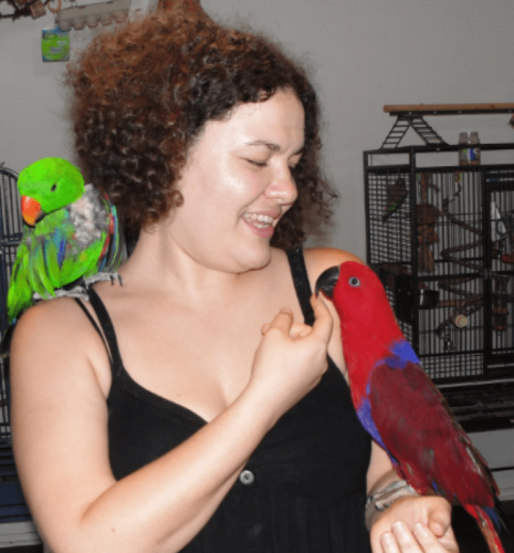 Image of a person with short, curly hair holding a red bird in their hand and a green bird on their shoulder.