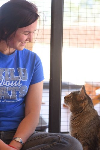 A woman sitting on the floor of a cat enclosure next to a tabby cat. They are looking at each other.