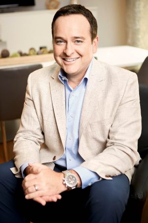 A white man with short brown hair seated with his hands folded.