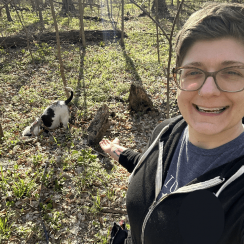 A woman with short hair standing in the woods with a dog in the background digging a hole.